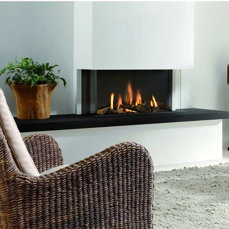 Element4 Trisore 100, tre-sidig gasspeis med rammeløst glass og realistiske flammer over keramiske vedkubber, plassert i en lys, minimalistisk stue med kurvstol, grønn plante og teksturert teppe